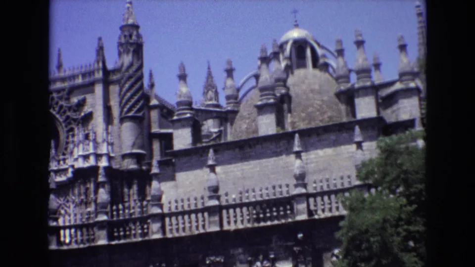 1972 Spain 8mm frame showing a wider view of the Gothic church exterior with spires, a dome, carved stonework, balcony railings, and trees near the base.