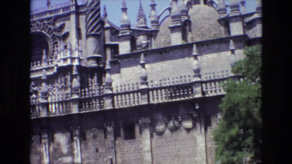 1972 Spain home-movie frame showing the church facade from street level, with Gothic stone details, spires, balcony railings, blue sky, and trees along the lower right.