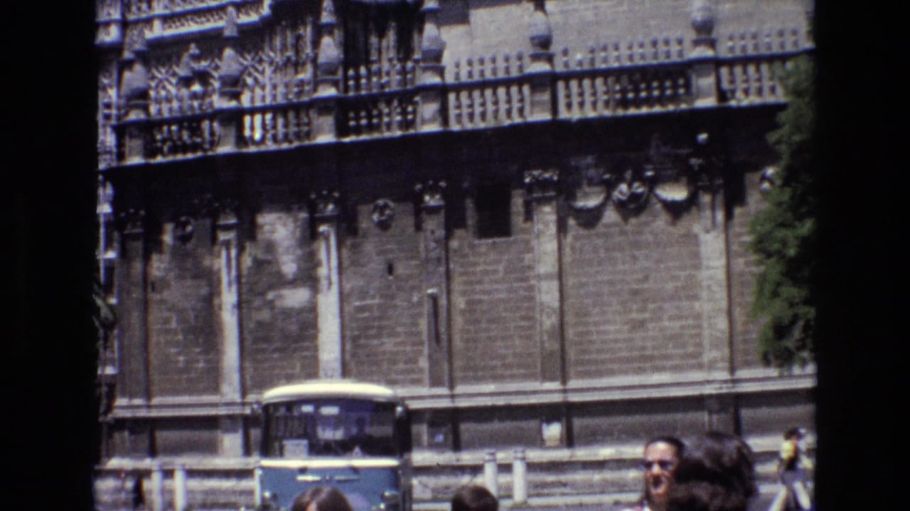 1972 Spain home-movie frame showing an ornate stone building near a street, with a blue-and-white bus and pedestrians partly visible in the foreground.