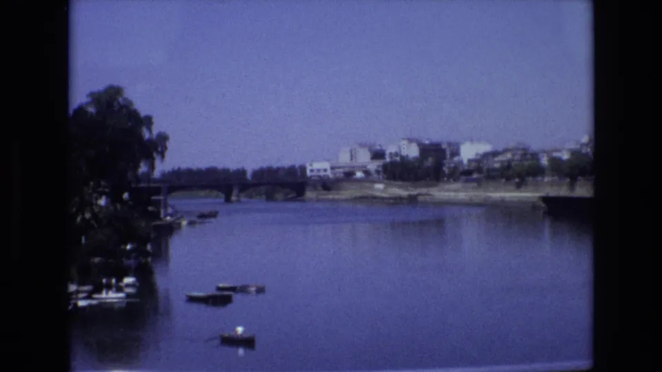 1972 Spain home-movie frame showing a calm wide river with small boats, a low bridge, trees along the bank, and distant riverside buildings.