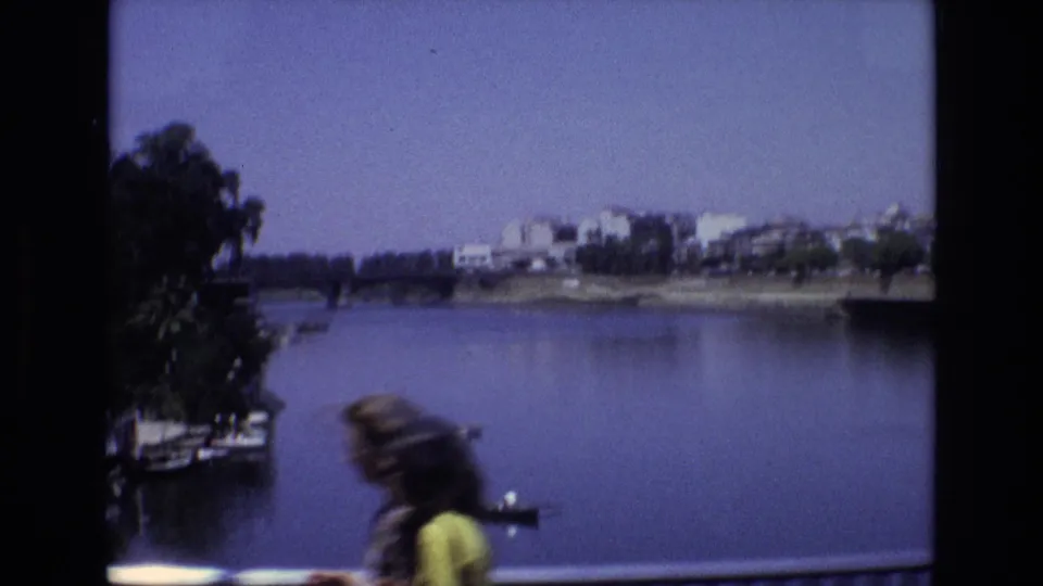 1972 Spain home-movie frame of the river, bridge, trees, and far-bank buildings, with a blurred pedestrian crossing the foreground near the railing.