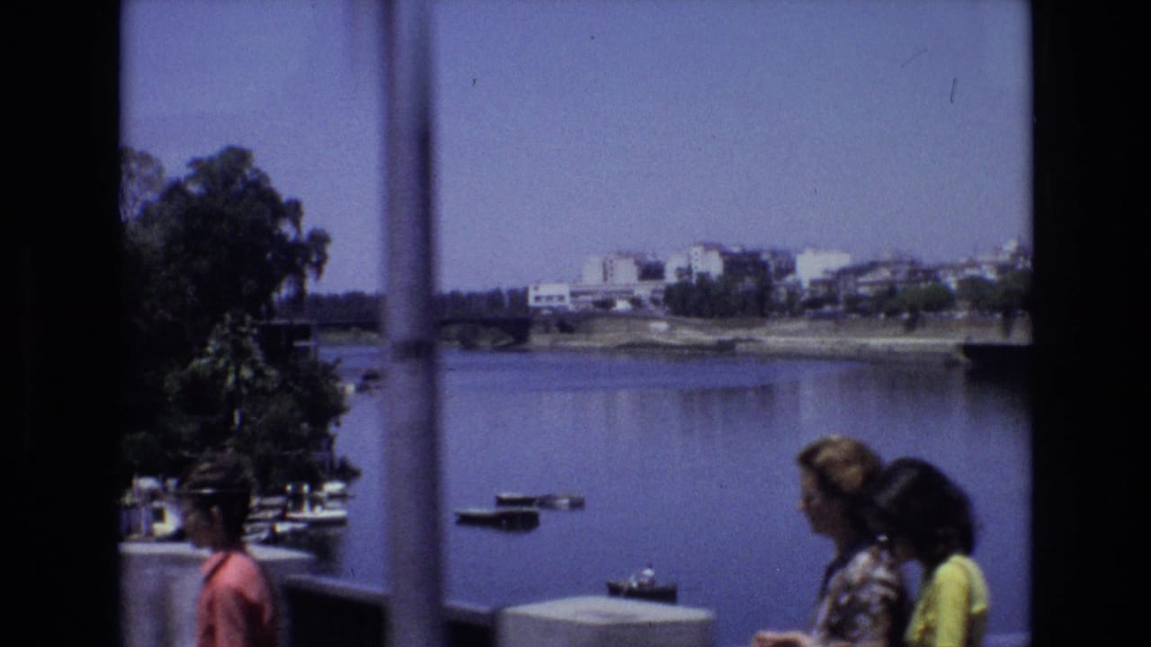 1972 Spain home-movie frame showing people walking beside a river, with trees, moored small boats, a distant bridge, and buildings across the water.