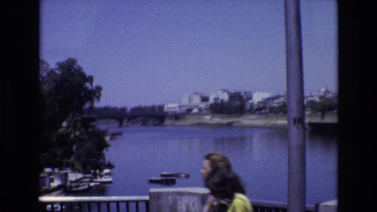 Two pedestrians walk past a railing above a river with trees, small boats, a bridge, and buildings in the distance.
