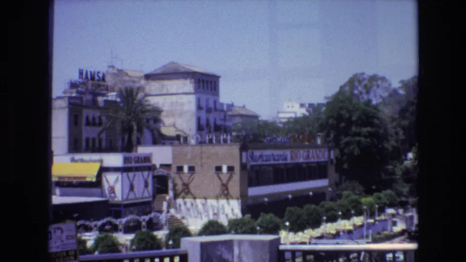 1972 Spain waterfront street scene showing restaurant and commercial signage, trees, yellow awnings or umbrellas, outdoor seating, and a foreground promenade railing.
