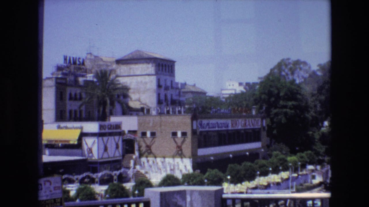 1972 Spain 8mm frame showing a sunlit urban square with Hamsa lettering, Restaurante Rio Grande signage, palm trees, and distant buildings.