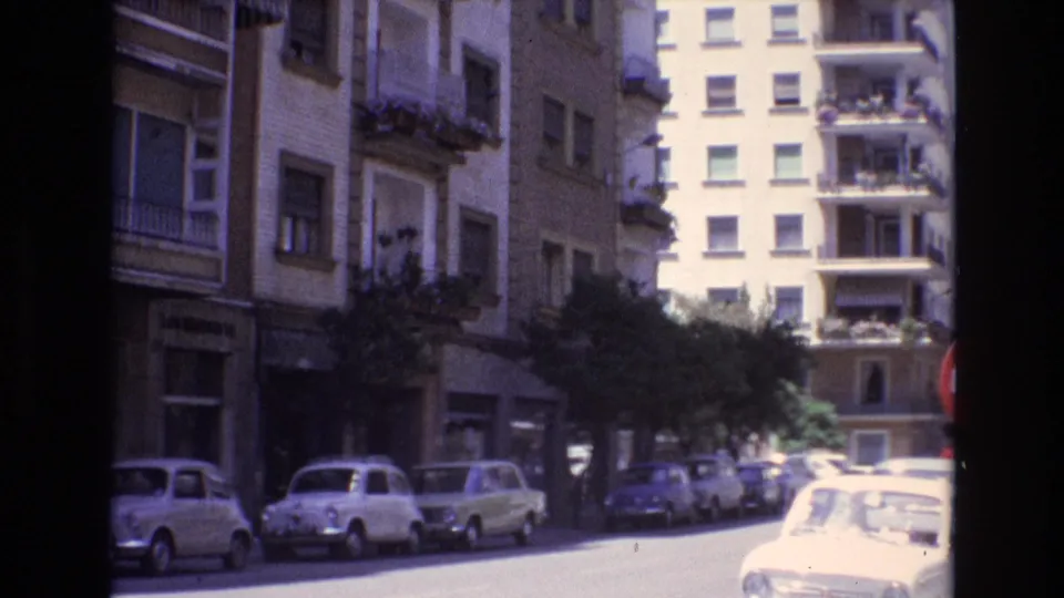 Spain 1972 frame looking down a residential street lined with apartment buildings, balconies, trees, and parked compact cars in bright sunlight.