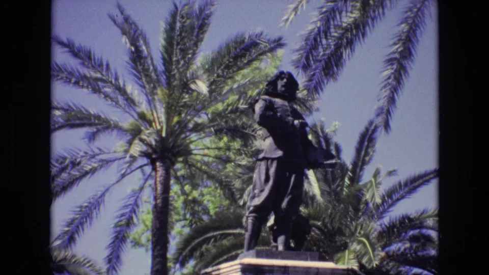 A dark civic statue on a pedestal is framed by palm fronds and clear sky in the supplied 1972 Spain 8mm travel footage.