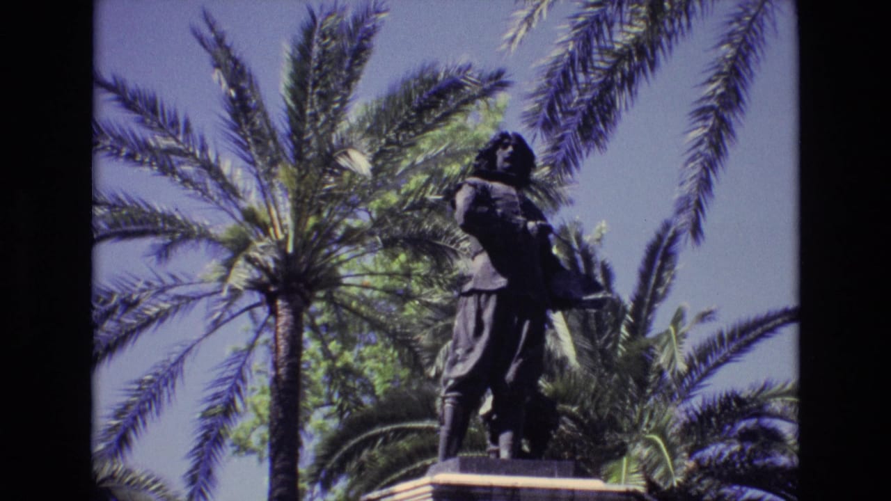 1972 Spain 8mm frame showing a dark bronze male statue on a pedestal, filmed from below with tall palms and blue sky behind it.