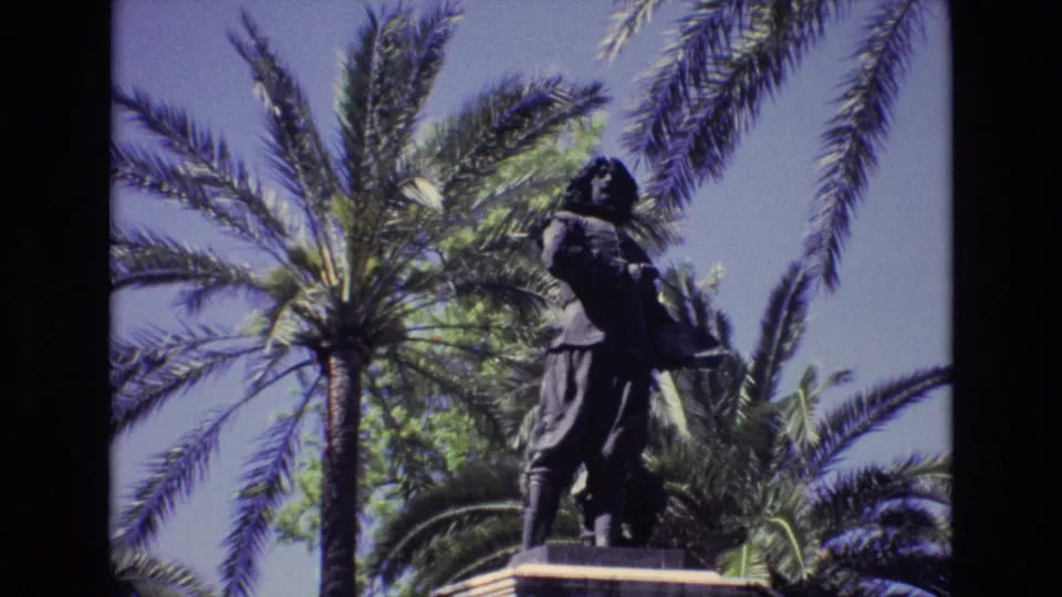 1972 Spain 8mm frame showing a dark bronze male statue on a pedestal, filmed from below with tall palms and blue sky behind it.