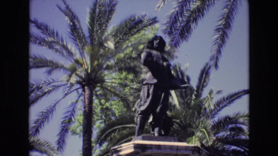 1972 Spain home-movie frame with the bronze statue slightly farther right in the composition, backed by palm trunks, green foliage, and clear sky.