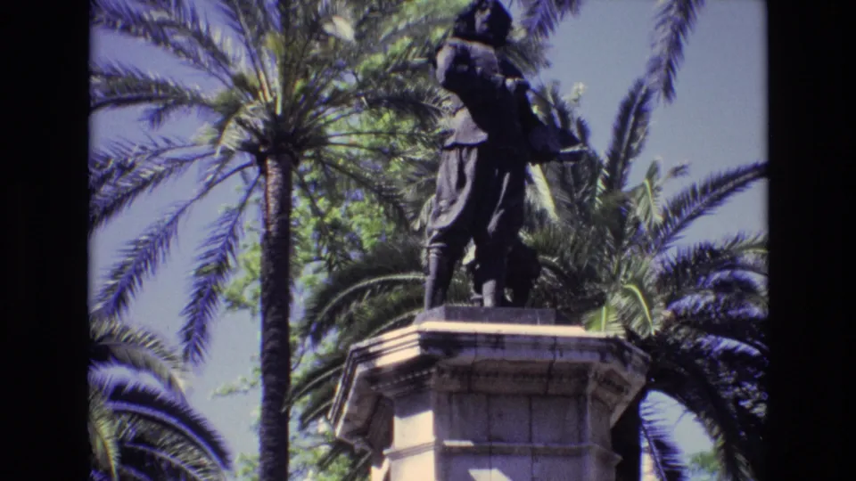 1972 Spain 8mm frame as the camera tilt reveals more of the stone pedestal beneath the bronze figure, with palms filling the background.