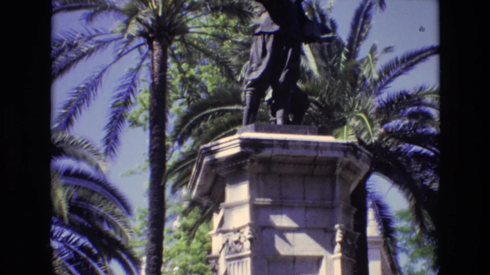 1972 Spain 8mm frame showing the bronze statue atop a larger visible stone base with decorative relief panels, surrounded by palm trees and sunlit sky.