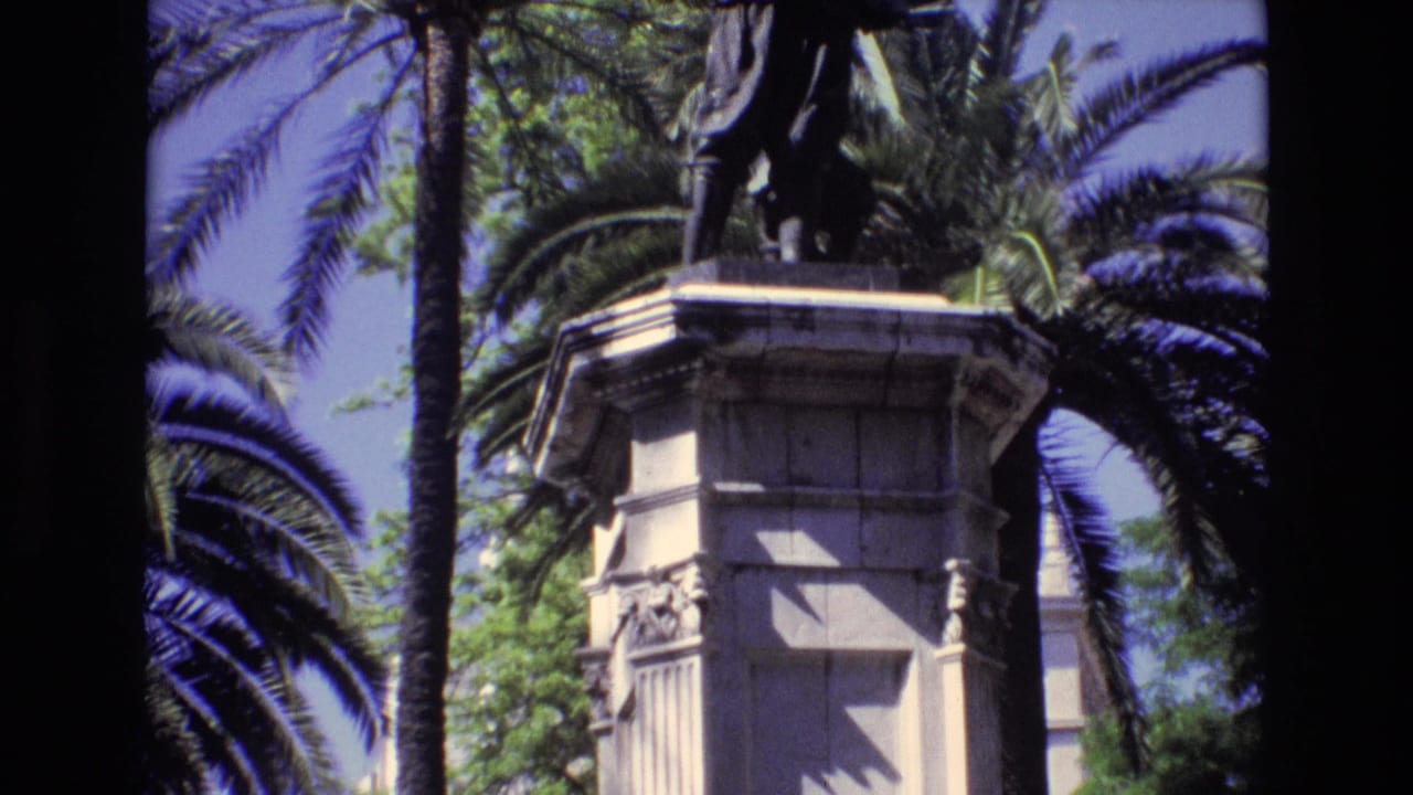 1972 Spain 8mm frame showing a close visitor view of a historic stone monument pedestal with palm trunks, palm fronds, leafy trees, and bright sky.
