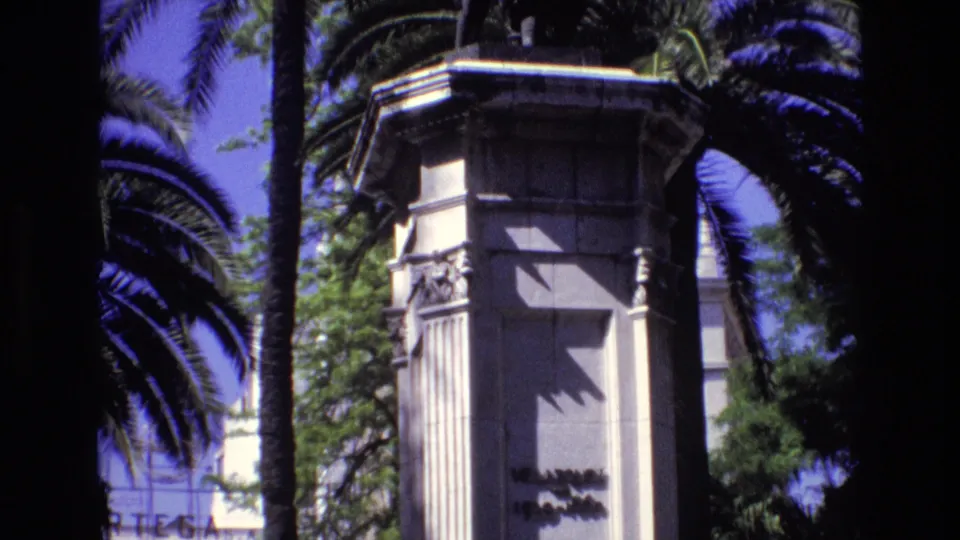 1972 Spain home-movie frame with the camera tilted lower on a stone monument pedestal, showing vertical carvings, palm trunks, foliage, and partial inscription shadowing.