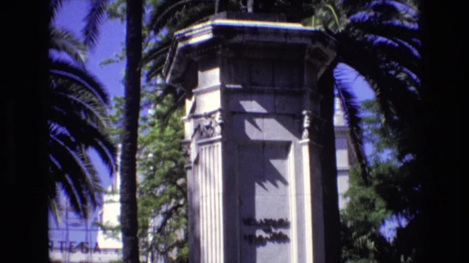 Final 1972 Spain 8mm frame holding on the pale stone monument pedestal, with carved side details, palm trunks, dark fronds, green trees, and sharp sunlight.
