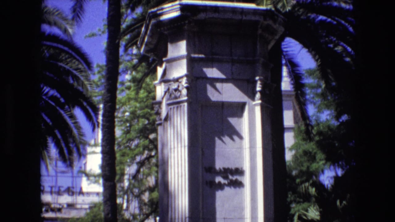 1972 Spain 8mm frame showing a tall white ornamental pillar with carved details, partial inscription, palm trunks, foliage, and blue sky.