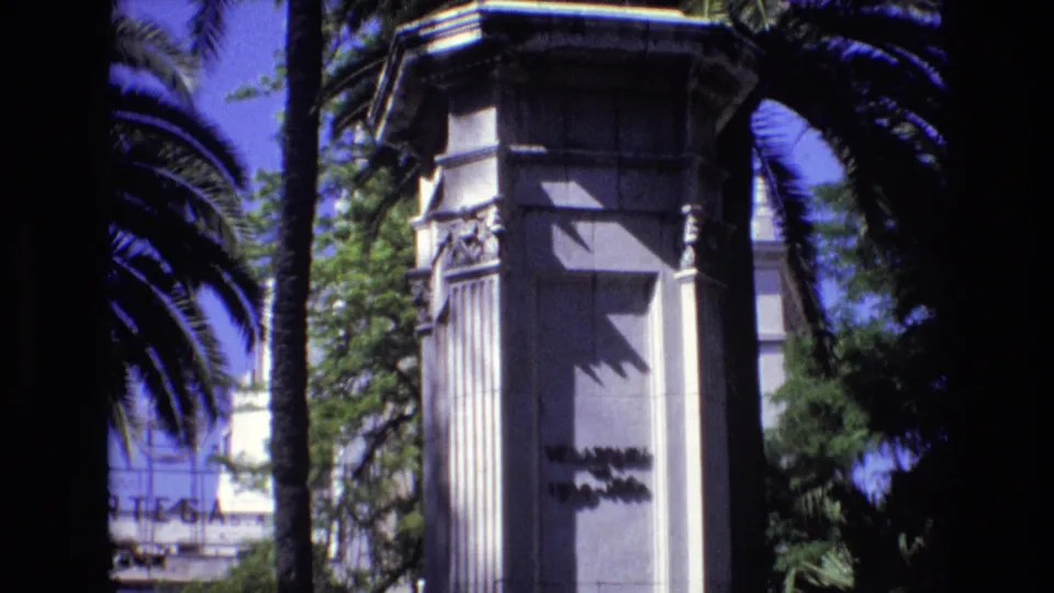 1972 Spain 8mm frame showing a tall white ornamental pillar with carved details, partial inscription, palm trunks, foliage, and blue sky.