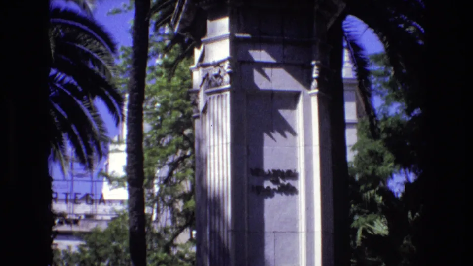 1972 Spain 8mm frame with a white stone pillar, dark palm trunk foreground, dense greenery, and partially visible lettering on a background structure.