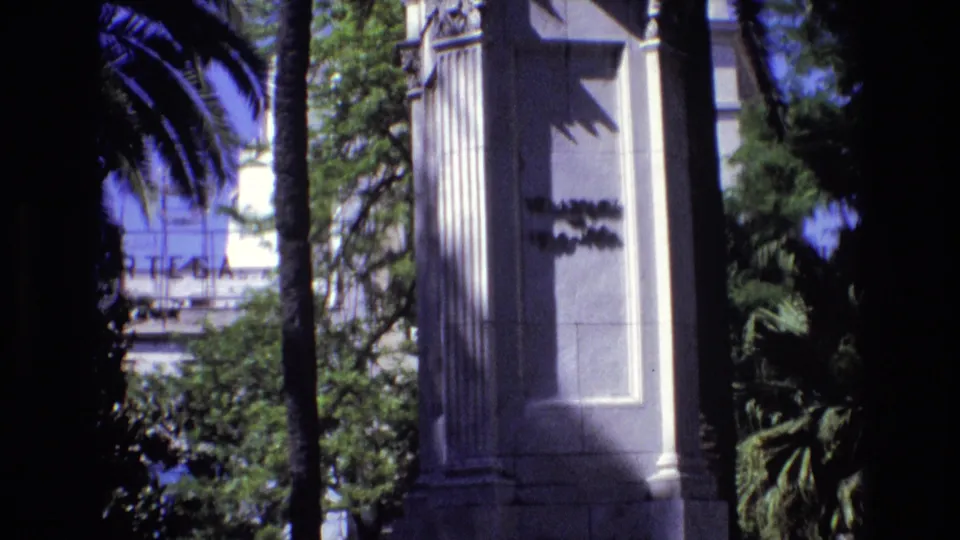 1972 Spain 8mm frame of an ornate white pillar with an unreadable partial inscription, palm trees, leafy vegetation, and bright sky behind it.