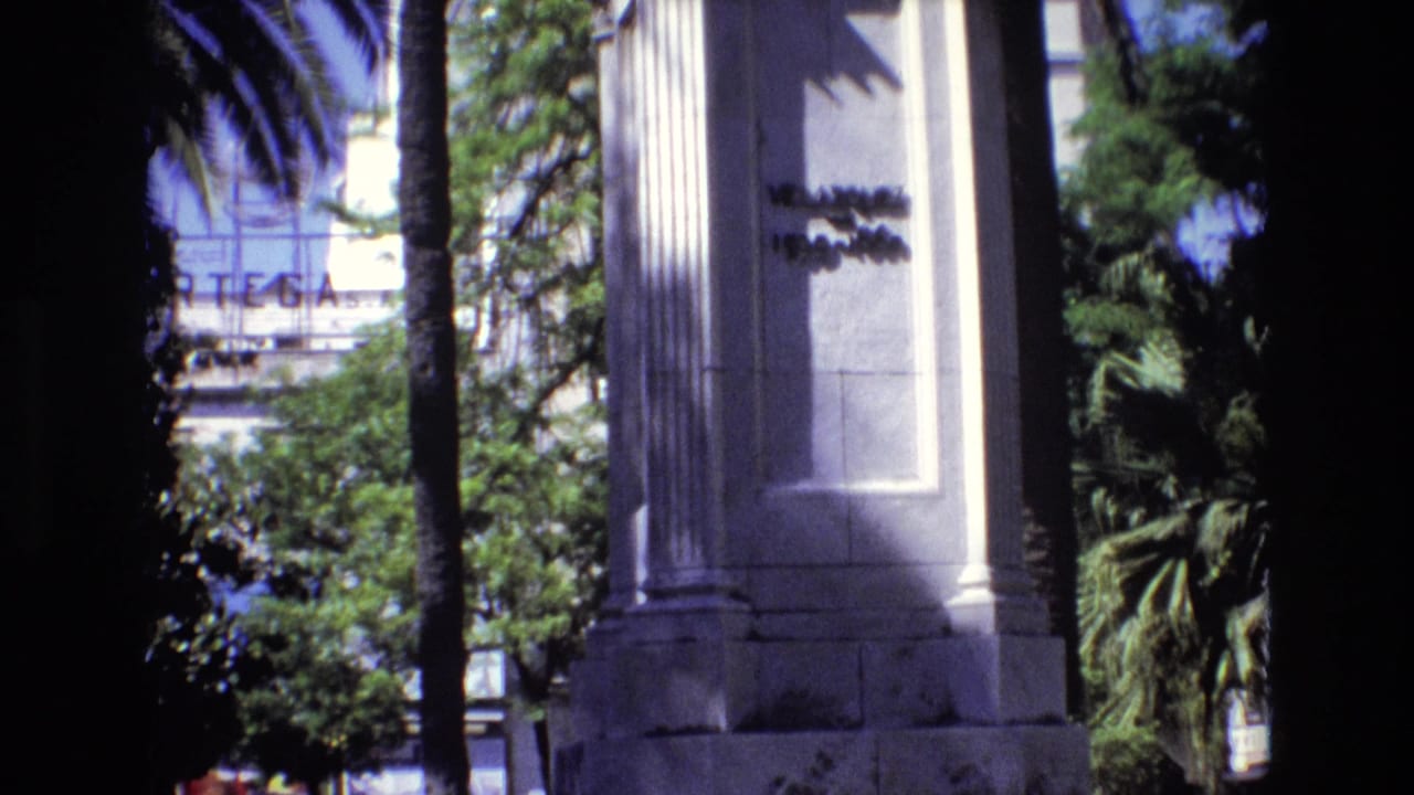 1972 Spain 8mm frame showing fluted stone monument columns, palm trunks, leafy landscaping, and a distant building with a partial sign.