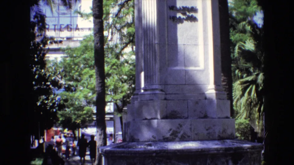 1972 Spain frame with the camera lower on the monument, showing weathered stone, palm trees, pedestrians on a sunlit sidewalk, and distant storefront signage.