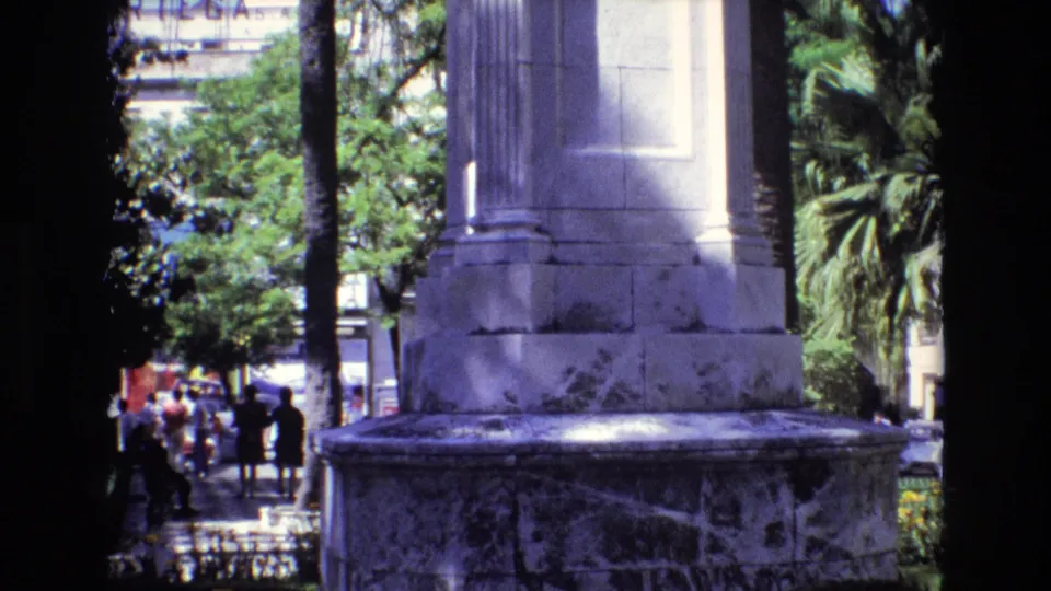 1972 Spain 8mm still showing the lower marble base of a public monument, palm-lined landscaping, yellow flowers, and pedestrians walking in the distance.