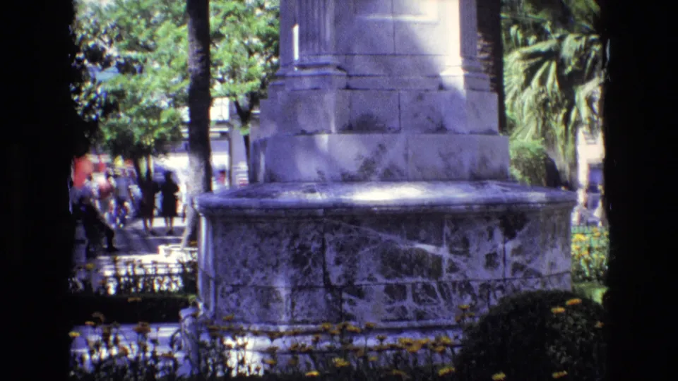 1972 Spain frame focused on the weathered stone base of the monument, with palms, flowerbeds, railings, and distant pedestrians in a public urban setting.