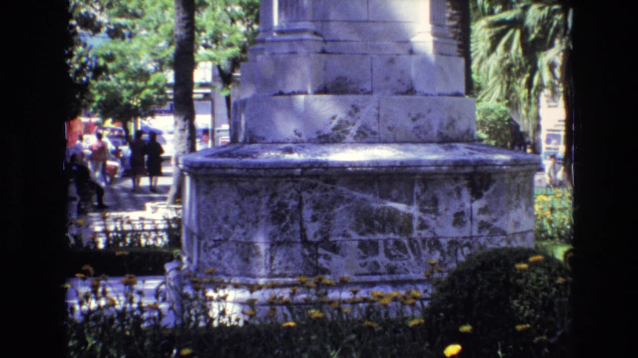 1972 Spain home-movie frame showing a large stone monument base bordered by yellow flowers, trees, and distant pedestrians in a sunny urban park.