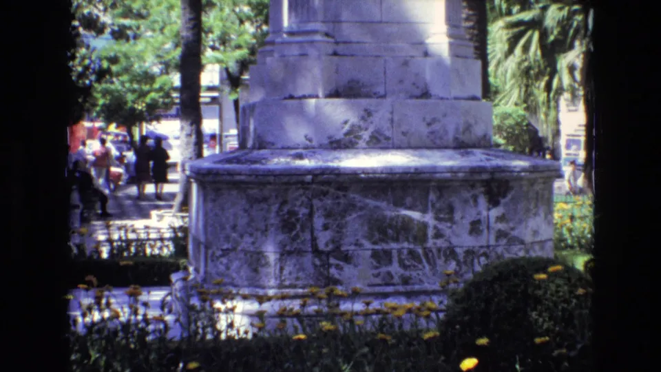 1972 Spain home-movie frame showing a large stone monument base bordered by yellow flowers, trees, and distant pedestrians in a sunny urban park.