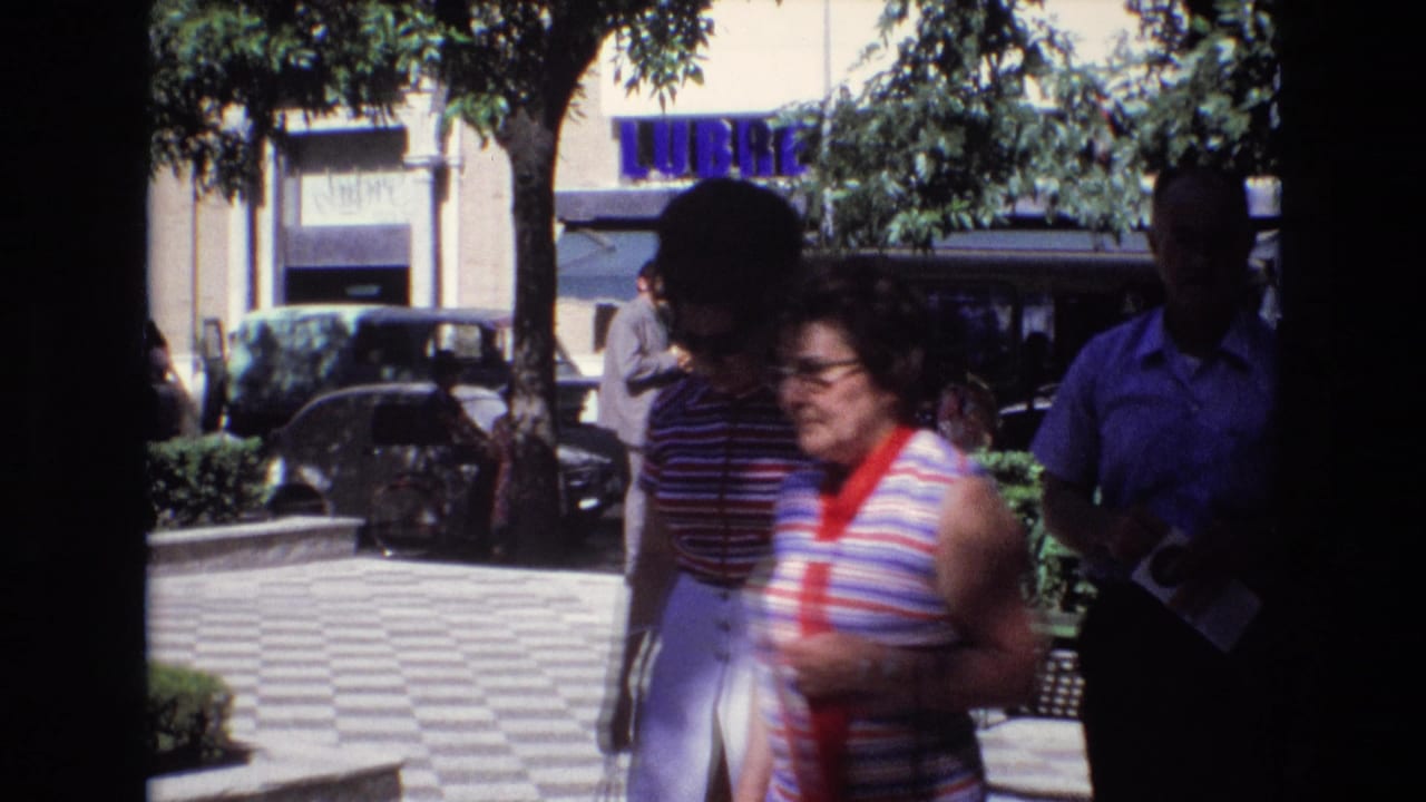Woman in a striped sleeveless top and red scarf walking past checkered pavement and trees.