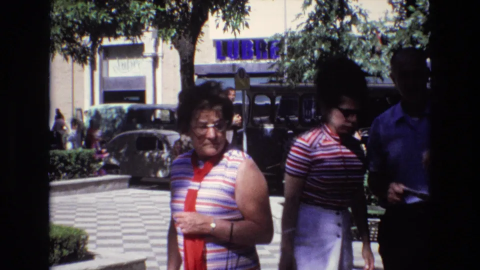 1972 Spain home-movie frame showing two women in striped tops and a man in a blue shirt walking through an urban park area with benches, trees, older vehicles, and a storefront sign behind them.