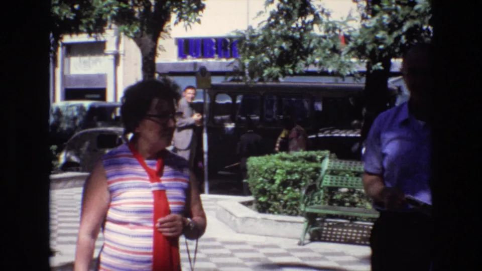 1972 Spain home-movie frame with a woman in a red scarf and striped sleeveless top walking past a green park bench, trees, hedges, parked cars, and a blue storefront sign.