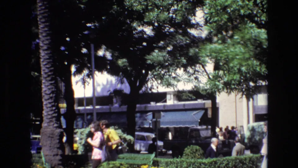 A wider 1972 Spain plaza view shows tall trees, green benches, pedestrians in warm-weather clothing, older cars, a bus, and building fronts seen from a handheld 8mm home-movie perspective.