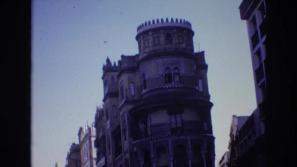 A handheld 1972 Spain street view frames an ornate rounded corner building with arched windows, balconies, and neighboring urban facades.