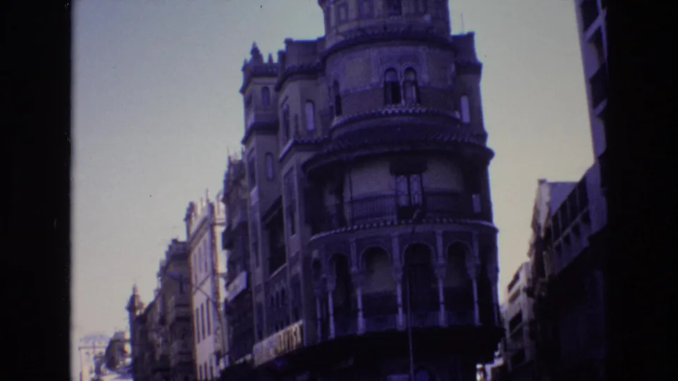 A 1972 Spain street frame shows the ornate corner building more fully, with arched lower openings, balcony railings, a streetlight, and nearby facades.