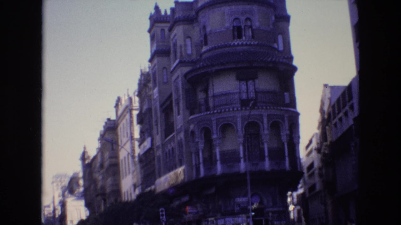 Spain 1972 home-movie frame showing an ornate rounded corner building with arched balcony openings above a city street with traffic and distant pedestrians.