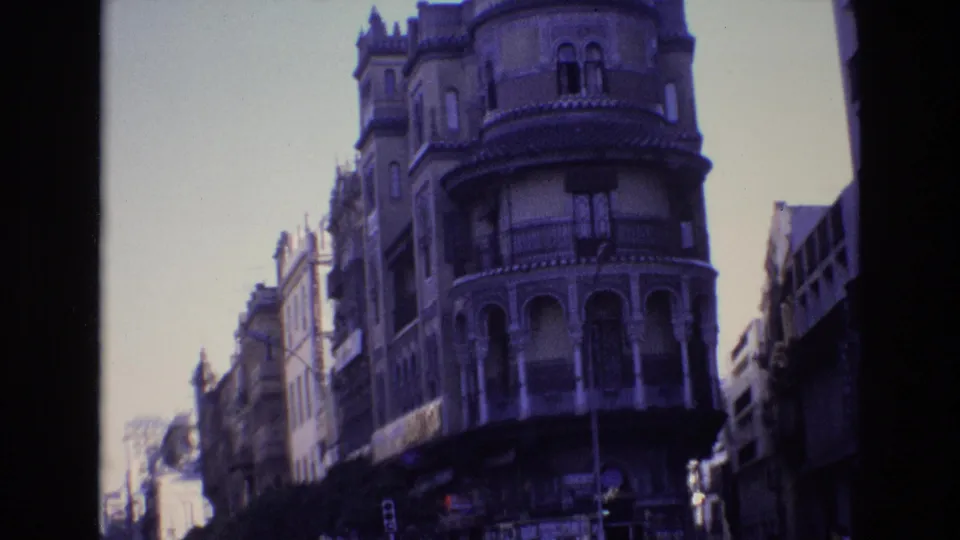 Spain 1972 home-movie frame showing an ornate rounded corner building with arched balcony openings above a city street with traffic and distant pedestrians.