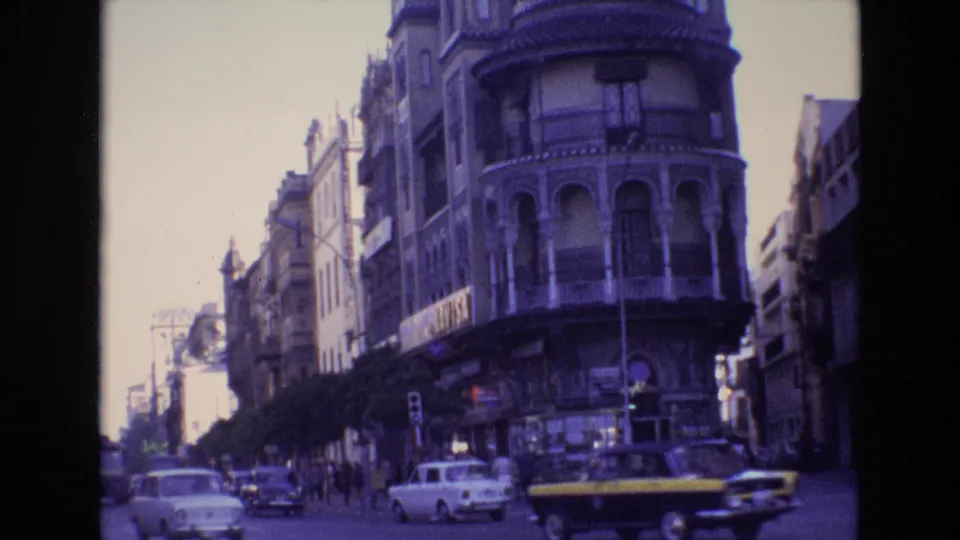 Spain 1972 home-movie still showing white sedans and a dark car moving past an ornate corner building with arched balcony details and pedestrians along the sidewalk.