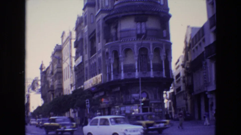 Spain 1972 frame with a white car, a yellow car, and other traffic passing beneath a rounded historic facade with arched openings and balconies.