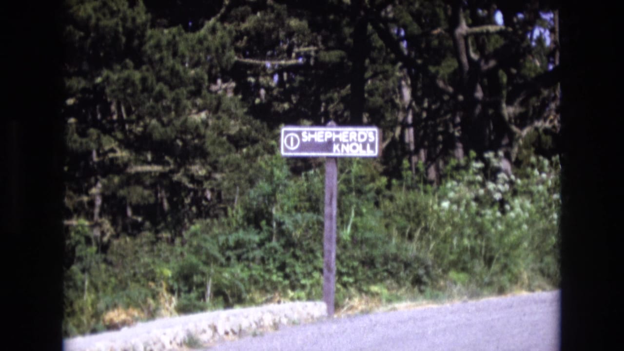 A Shepherd's Knoll roadside sign stands beside a paved edge with dense greenery in metadata-supported 1973 Central Coast California home-movie footage.