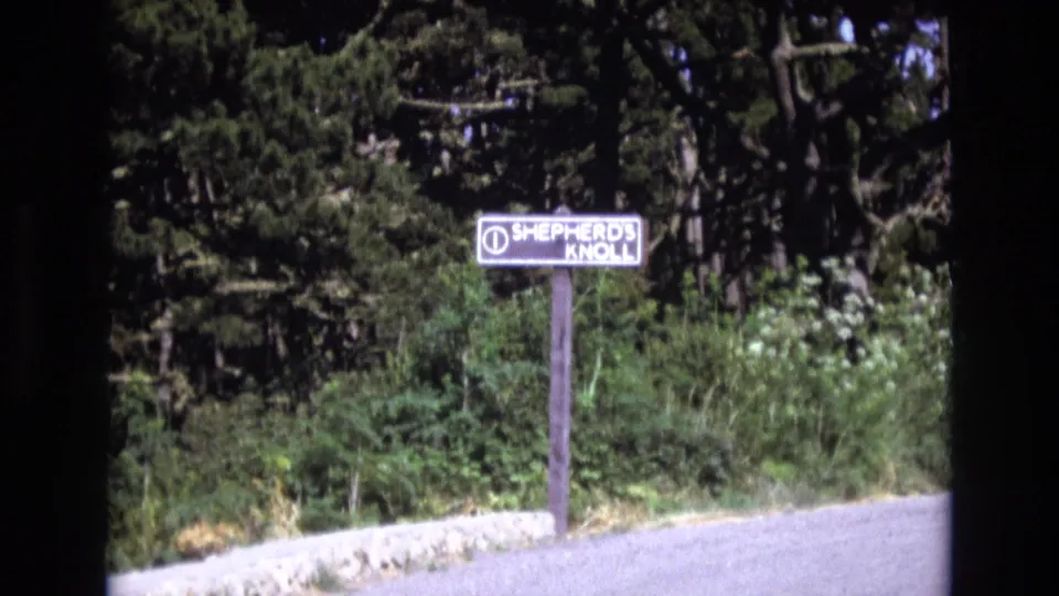 A Shepherd's Knoll roadside sign stands beside a paved edge with dense greenery in metadata-supported 1973 Central Coast California home-movie footage.