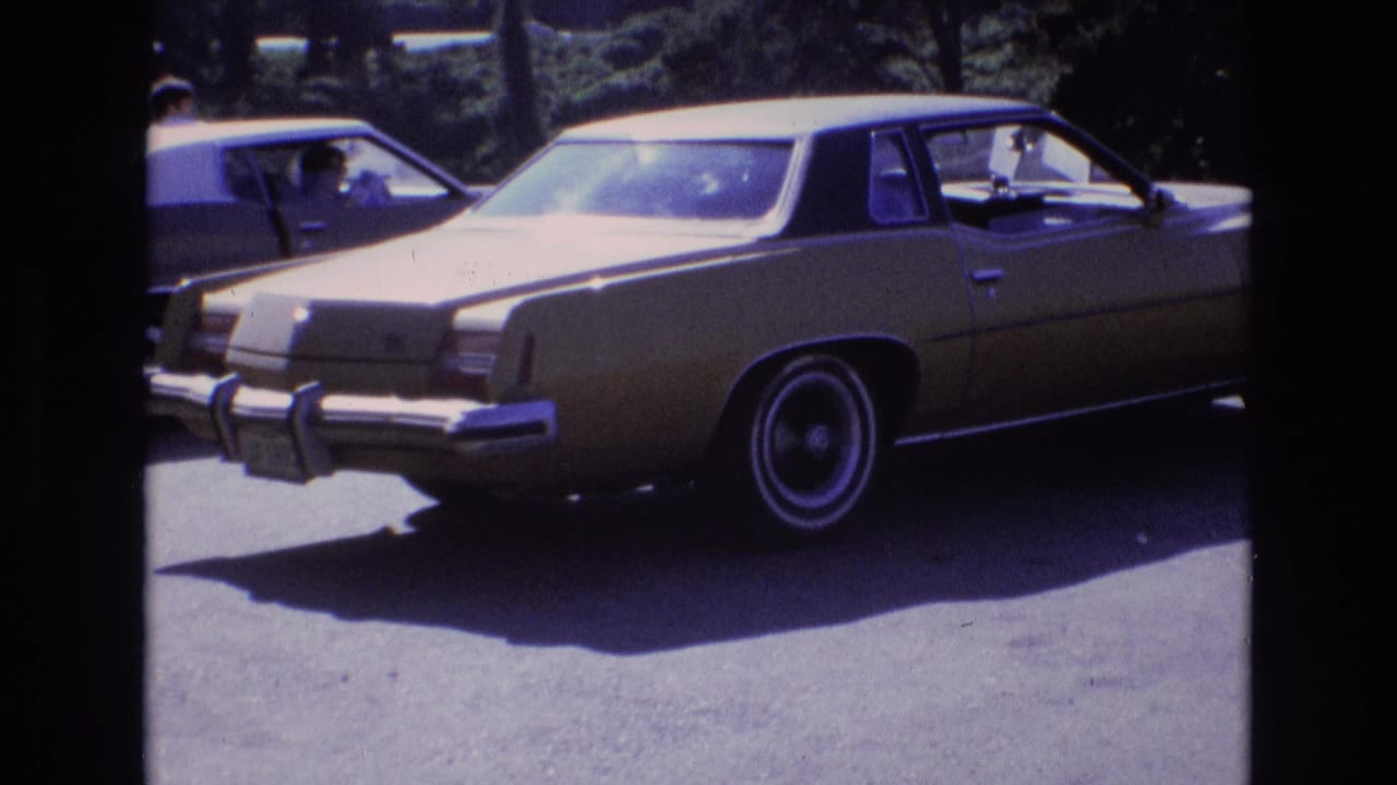 A gold-colored 1970s-style sedan with chrome bumper and whitewall tires is parked in a sunny lot, with another vehicle and a person visible in the background area.
