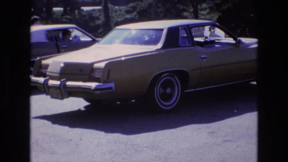 A gold-colored 1970s-style sedan with chrome bumper and whitewall tires is parked in a sunny lot, with another vehicle and a person visible in the background area.