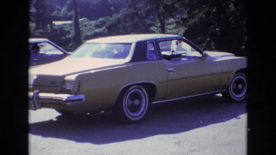A gold-colored sedan remains parked in bright sunlight against trees and a paved lot, with the 8mm frame edge visible around the scene.