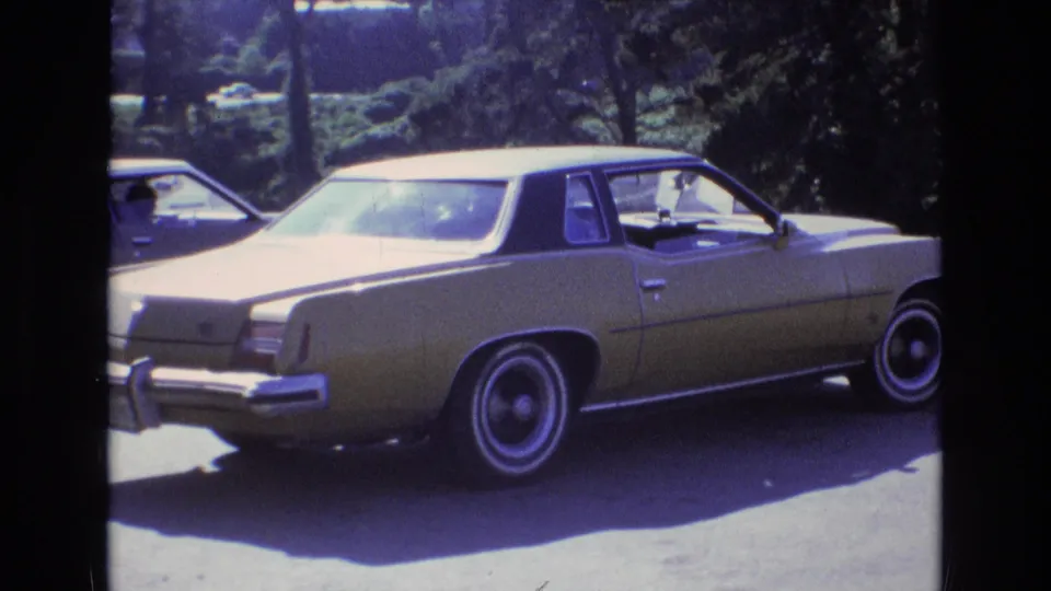 A gold 1970s-style American sedan with a dark roof and chrome bumper sits on a sunlit asphalt lot in Central Coast California footage from 1973.