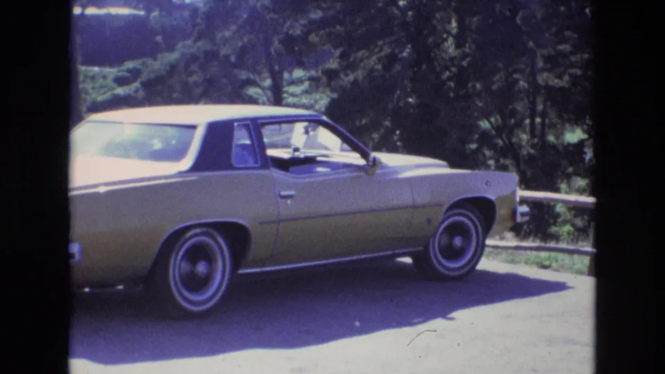 A closer 8mm frame shows the gold classic sedan cropped at the rear and front, with the open window area, whitewall tire, asphalt, and foliage visible.
