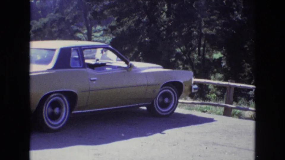 The final frame holds on the gold 1970s American sedan parked on asphalt near a wooden fence, with dense green trees behind the car.