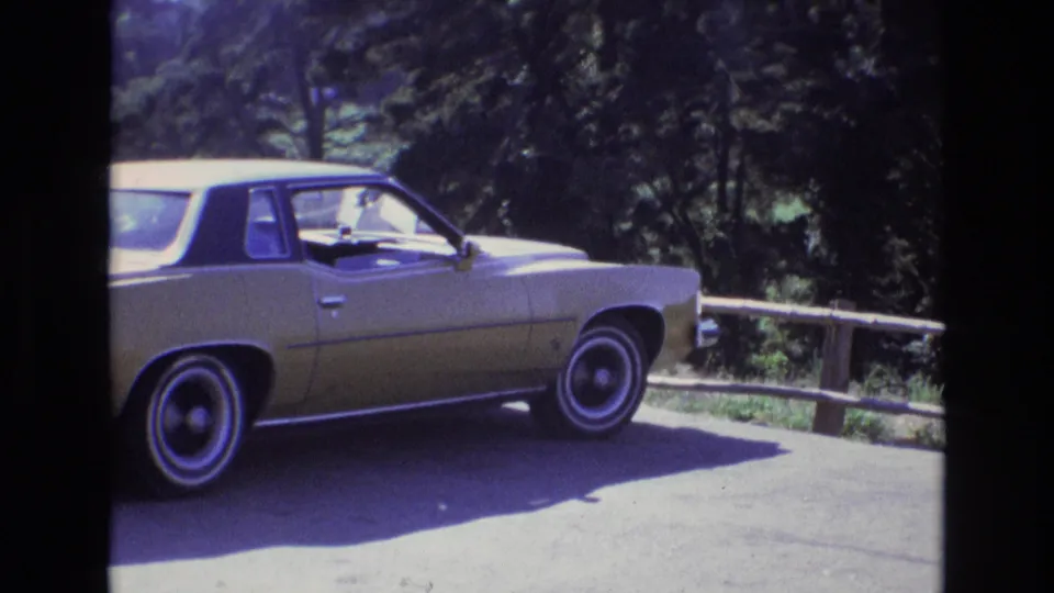 A yellow and black early-1970s-style car sits on a paved roadside overlook in coastal California, with a rustic log rail and dense trees behind it.