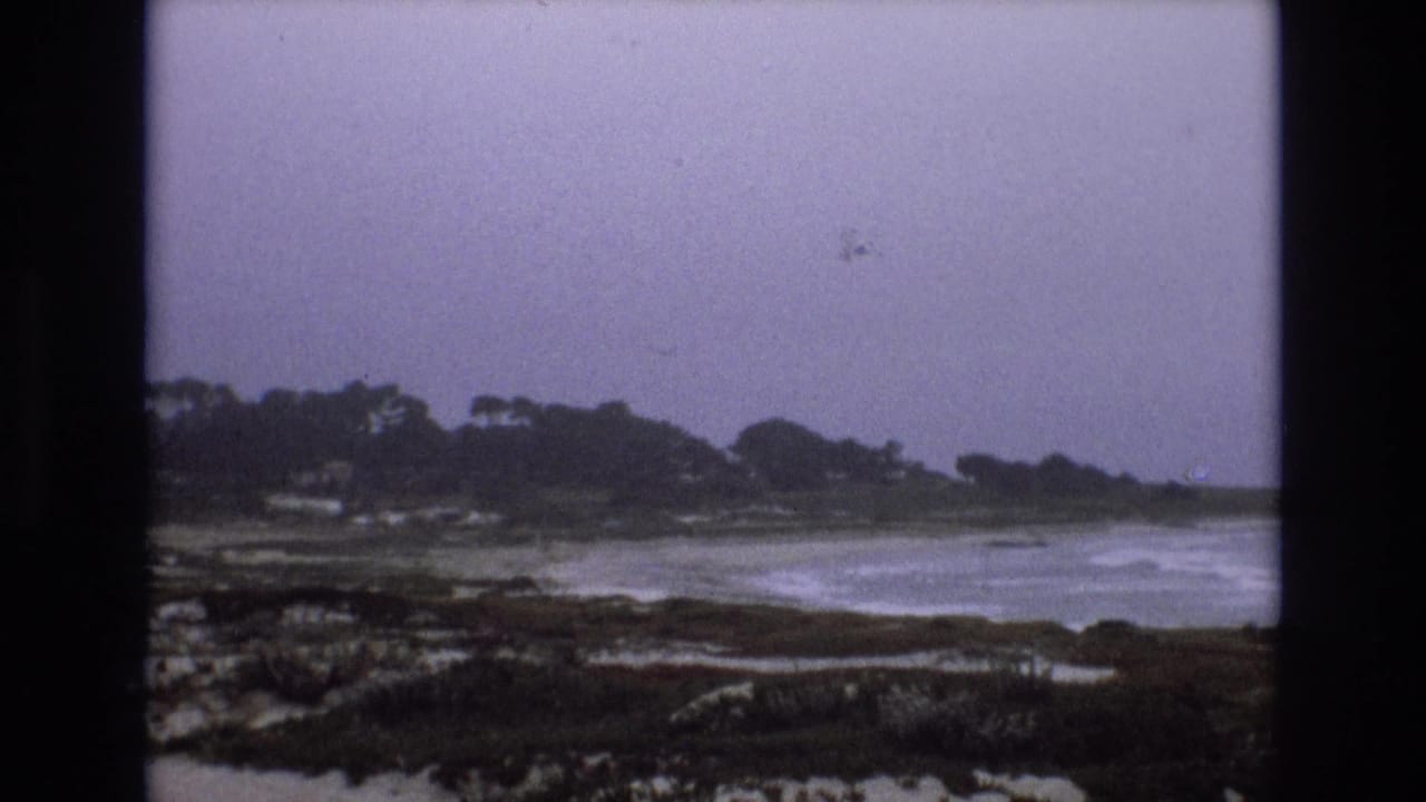 1973 8mm frame showing muted surf along a rocky California coast, with low vegetation, distant trees, and an overcast sky.
