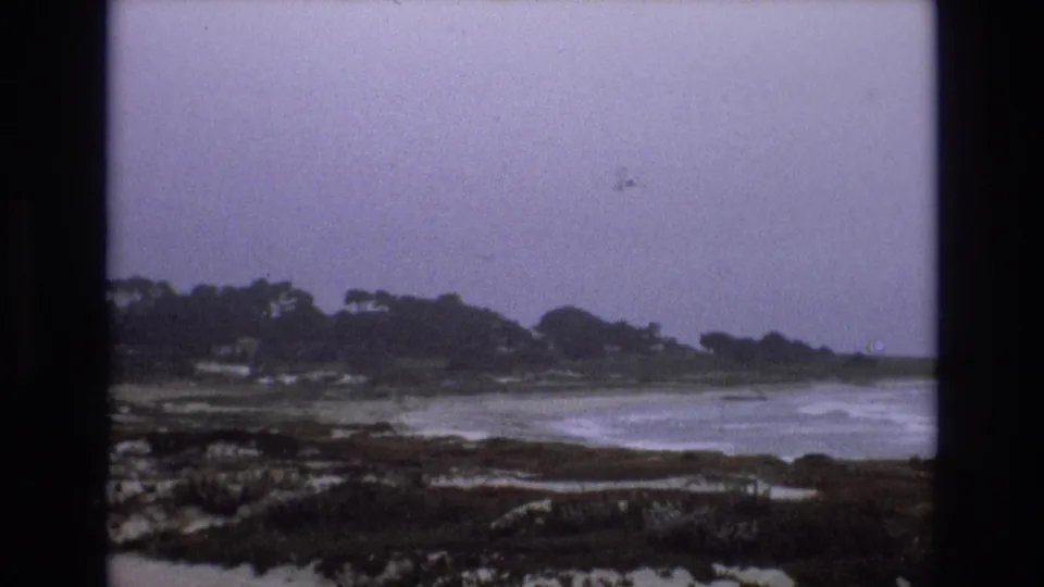 1973 8mm frame showing muted surf along a rocky California coast, with low vegetation, distant trees, and an overcast sky.